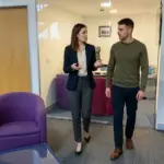 Two colleagues walk through a UK office discussing email marketing budget planning.
