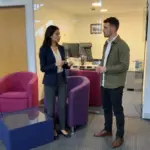 Two colleagues discuss Shopify project scope in an office while one makes a restrained pricing gesture.