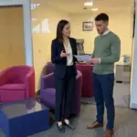 Two colleagues discuss a publicity plan in an office while reviewing an unreadable briefing sheet.
