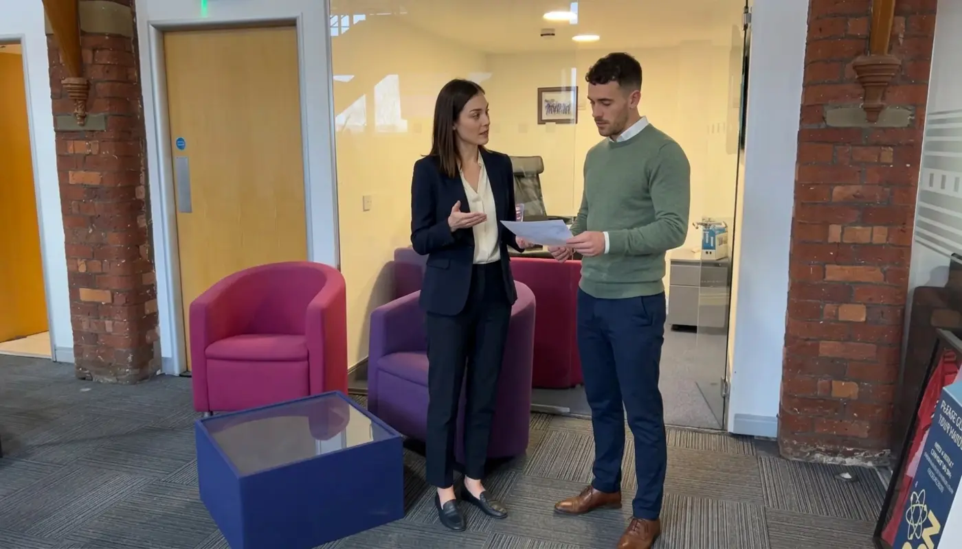 Two colleagues discuss a publicity plan in an office while reviewing an unreadable briefing sheet.