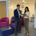 Two colleagues discuss an ecommerce launch plan inside a UK office while reviewing an unreadable tablet.