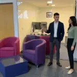 Two colleagues discuss CRM workflow in a UK office while standing in open floor space.