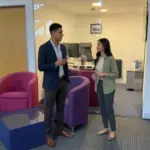 Two colleagues discuss an AI research workflow in a UK office while reviewing a shared work area.