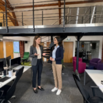 Two colleagues review a workstation together in a modern office while discussing an email marketing plan.