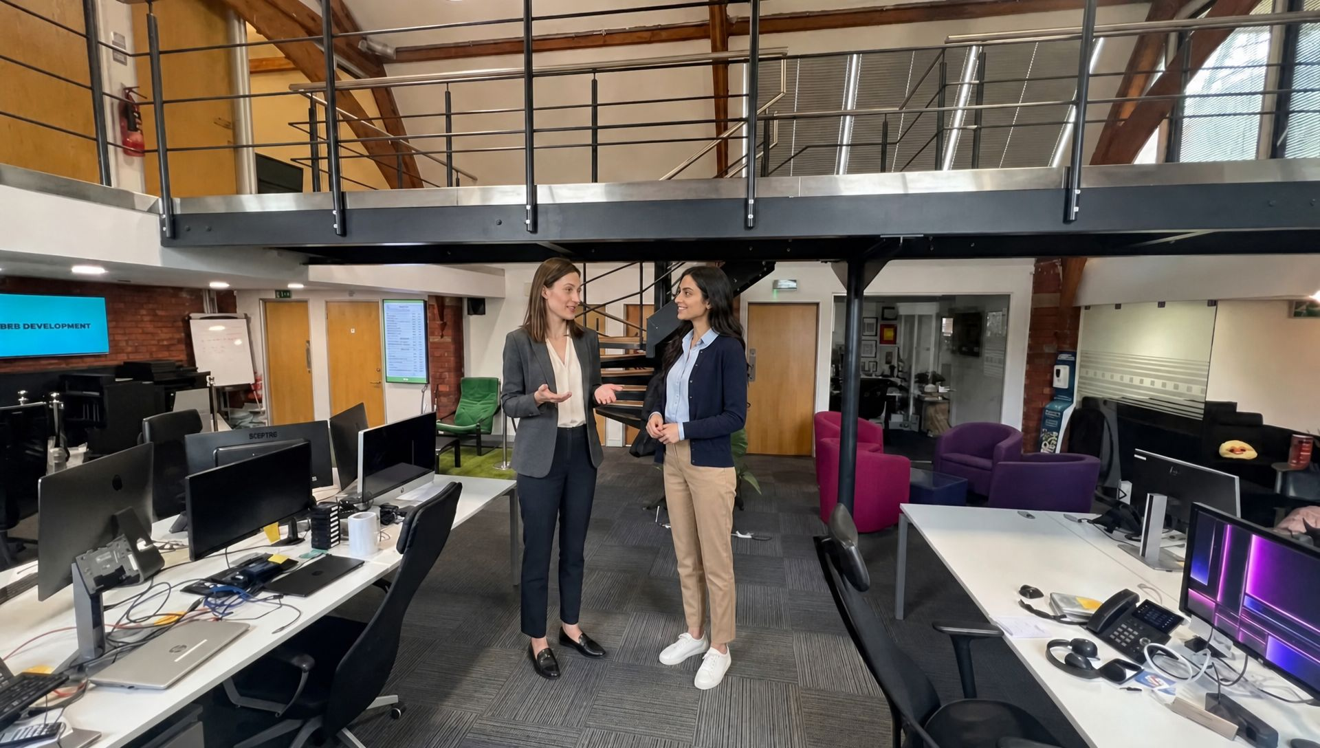 Two colleagues review a workstation together in a modern office while discussing an email marketing plan.