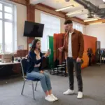 Two colleagues in an office review a phone notification setting beside a blue chair
