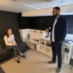 Two colleagues discuss brand lettering in a small office, with one seated on the sofa and one standing nearby.